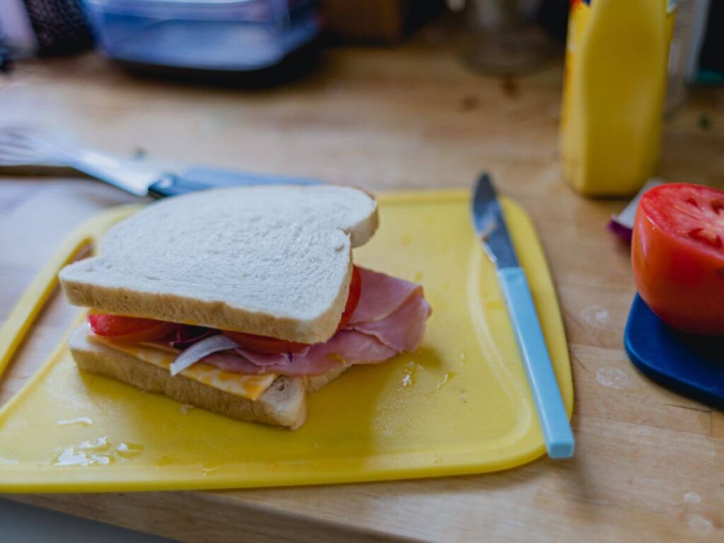 a freshly prepared ham sandwich on a yellow plastic chopping board, condiments and utensils around