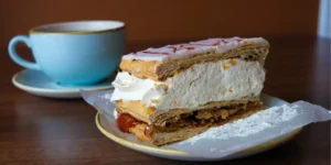 Slice of cream mille-feuille cake on plate with cup of coffee in background